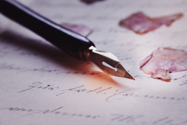 Närbild på gammaldags penna liggande på ett papper med text skrivet i skrivstil. På pappret ligger även enstaka rosenblad.